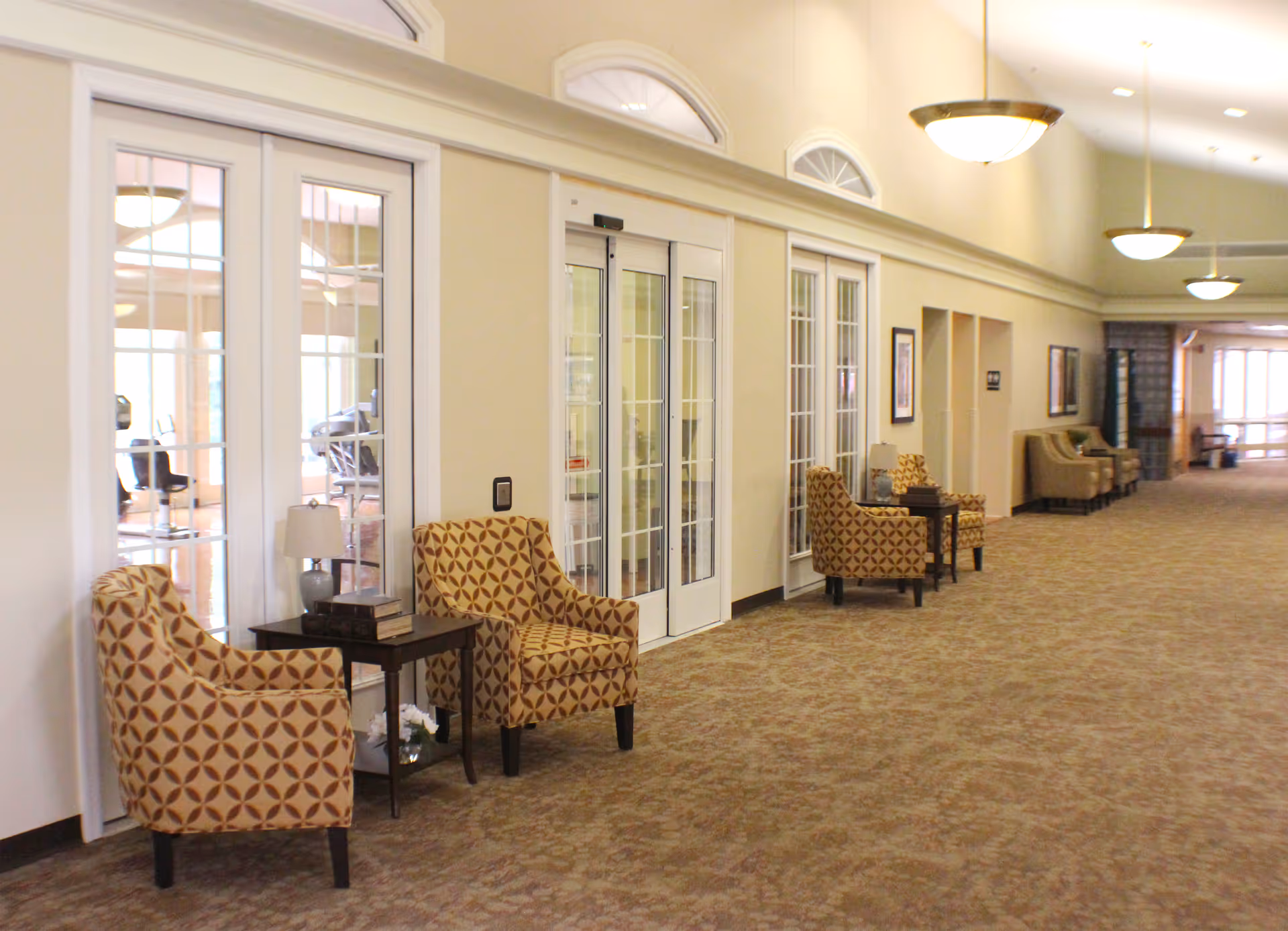 A long, well-lit hallway in a senior living facility with patterned armchairs and small tables with lamps placed along the wall. The hallway has large glass doors and windows on one side, and framed artwork on the walls. The carpet is patterned, and ceiling lights hang down providing warm lighting.