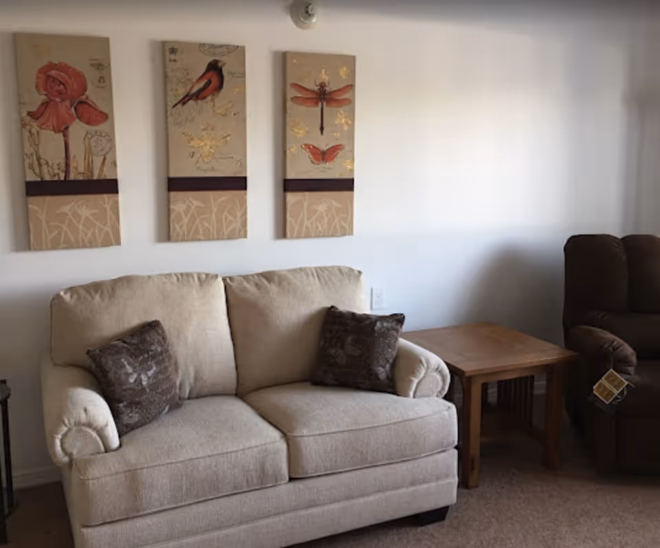 A cozy living room area with a beige loveseat featuring two dark patterned throw pillows, a wooden side table, and a brown recliner chair. Above the loveseat, three vertical decorative wall art pieces depict a flower, a bird, and insects on a beige background.