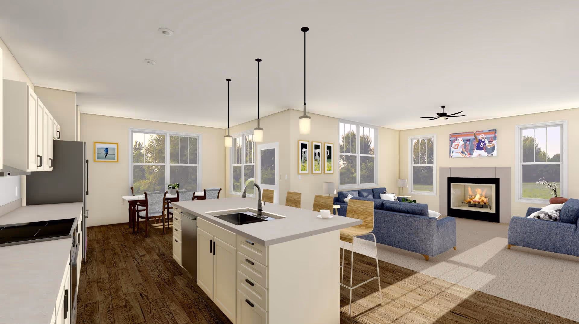 A bright open-concept interior space featuring a modern kitchen with white cabinetry, a large island with a sink and three pendant lights above it, and wooden flooring. Adjacent to the kitchen is a dining area with a table and chairs near large windows. The living room area includes blue sofas, a fireplace with a fire burning, a ceiling fan, and a wall-mounted TV showing a football game. The walls are light-colored with framed pictures and large windows letting in natural light.