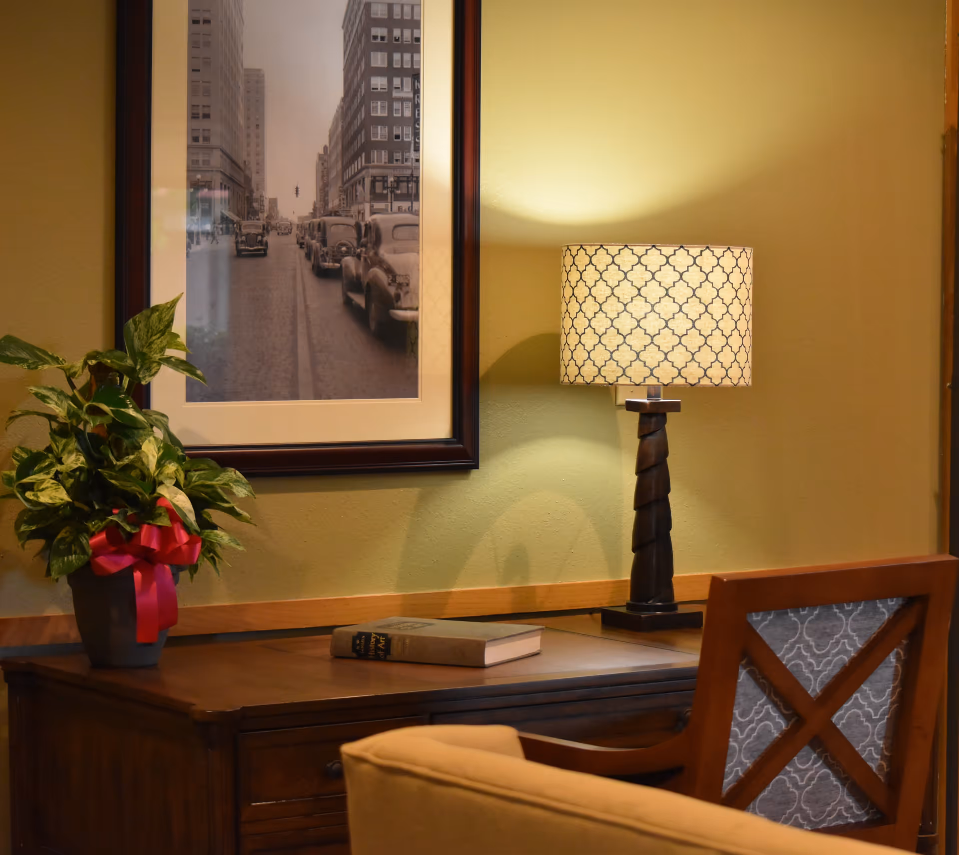 A cozy interior corner with a wooden desk, patterned table lamp, framed black-and-white city photo, potted plant, and chair.