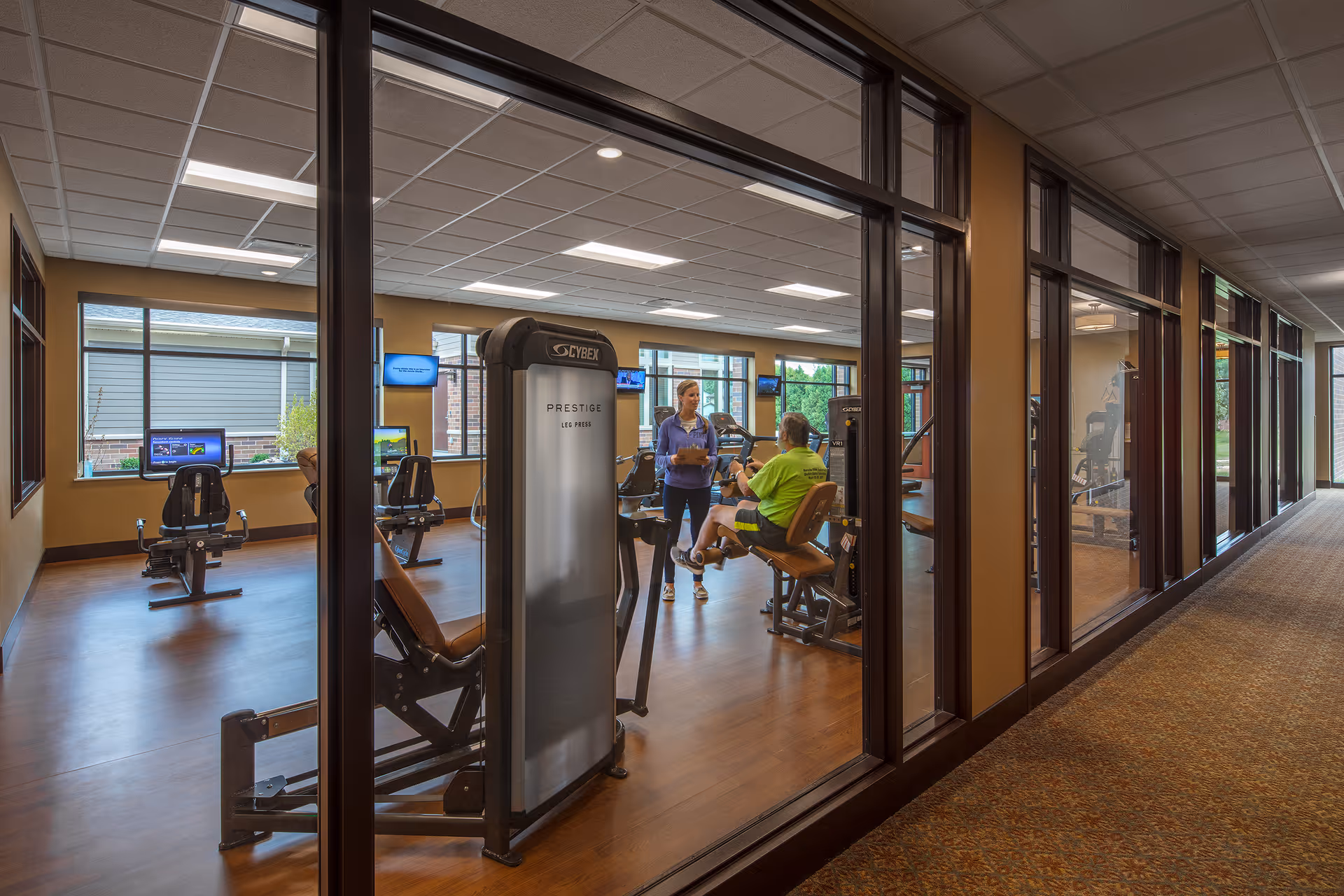 Glass-walled fitness room with exercise machines and two people talking inside.
