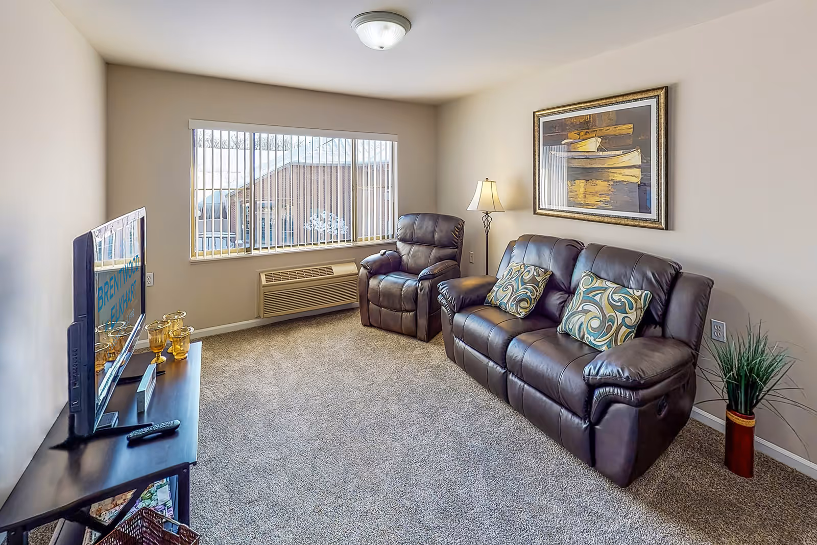 A cozy living room with beige walls and carpeted floor featuring a dark brown leather sofa and matching recliner chair, both adorned with decorative pillows. A floor lamp stands beside the sofa, and a framed painting of boats hangs on the wall above it. A flat-screen TV is placed on a black TV stand opposite the seating area, with a window behind the recliner letting in natural light and showing a view of a building outside.