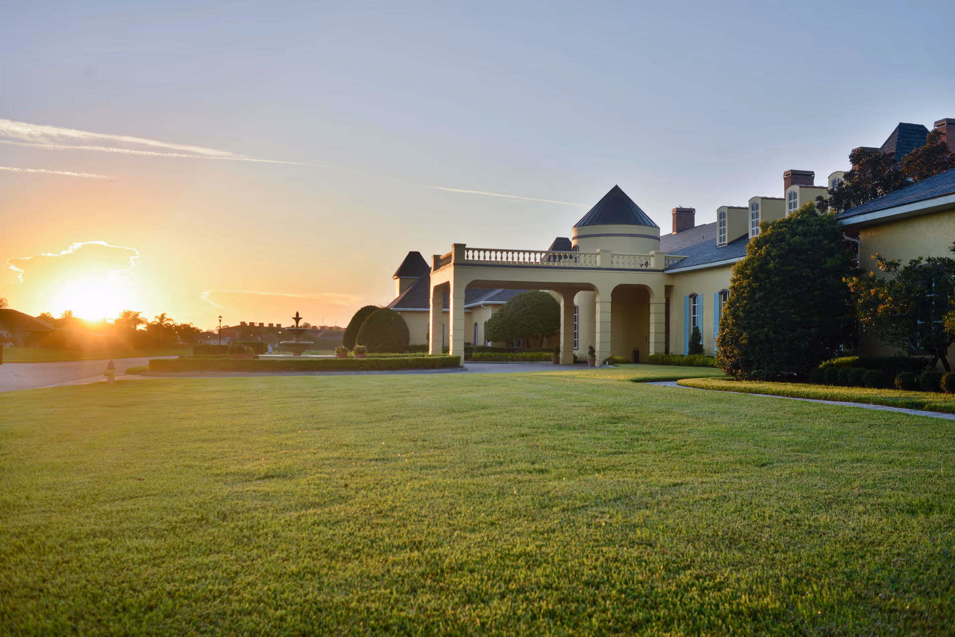 Sunset view of a large yellow building front with a covered portico, trimmed hedges, and an expansive green lawn.