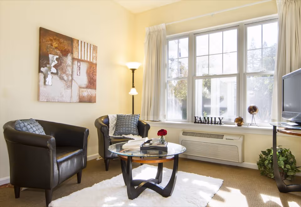 A cozy living room with two black leather armchairs, a round glass coffee table with books and a small vase with a red flower, a floor lamp, a large window with white curtains, and a decorative sign that says FAMILY on the windowsill. There is a TV on a stand and a green plant in the corner.