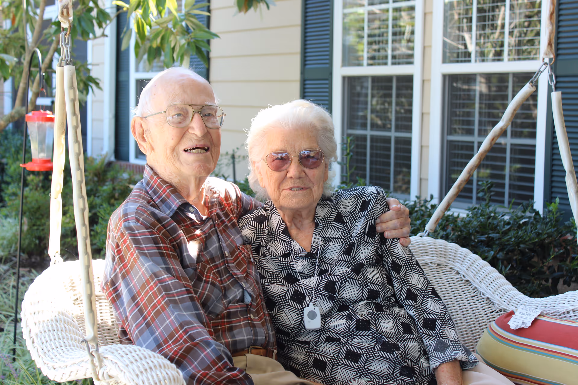 An elderly man and woman sitting closely together on a white wicker porch swing outside a building with beige siding and green shutters. The man is wearing glasses and a plaid shirt, while the woman is wearing sunglasses and a patterned blouse with a medical alert pendant. There are plants and windows in the background.