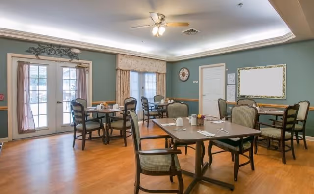 A dining room in a senior living facility with several tables and chairs arranged neatly. The room has wooden flooring, green walls with a wooden trim, and large windows with curtains allowing natural light to enter. A ceiling fan with lights is mounted on the ceiling, and there is a clock and a framed board on one wall.