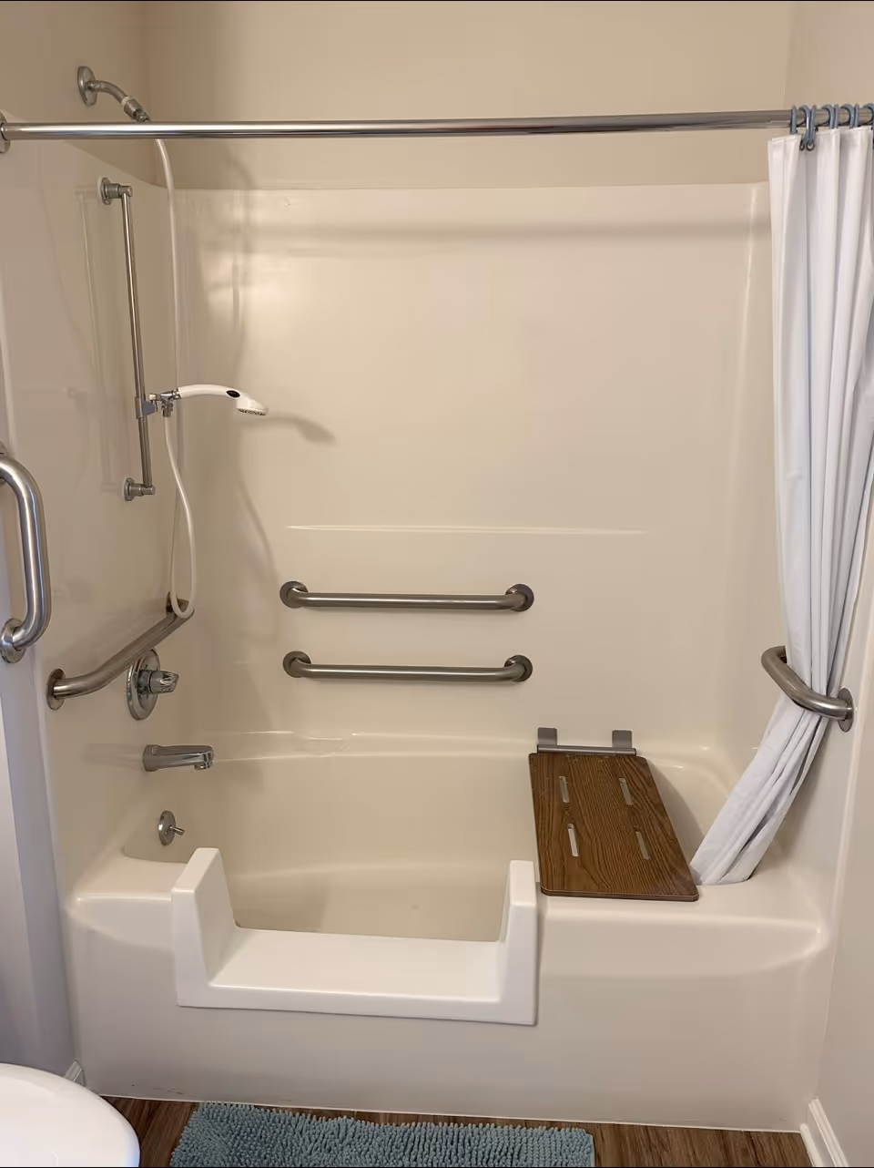A bathroom shower area with a built-in seat, grab bars on the walls, a handheld showerhead, and a white shower curtain pulled to one side. The shower has a lowered entry for accessibility and a wooden seat attached to the tub edge.