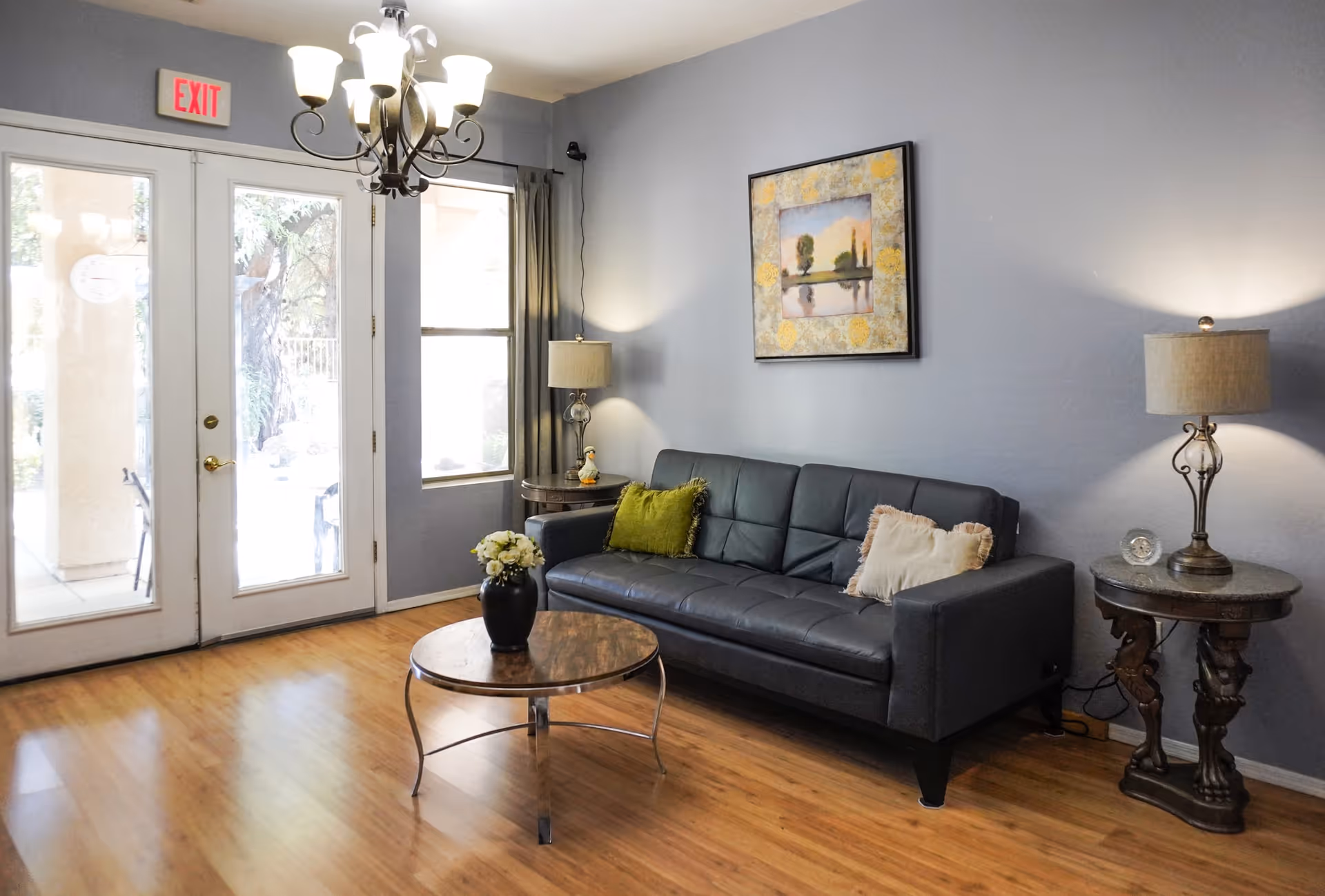 A cozy living room with a black leather sofa adorned with two decorative pillows, a round wooden coffee table with a vase of flowers, two side tables each with a lamp, a framed painting on the wall, and a glass door with an exit sign above it letting in natural light.