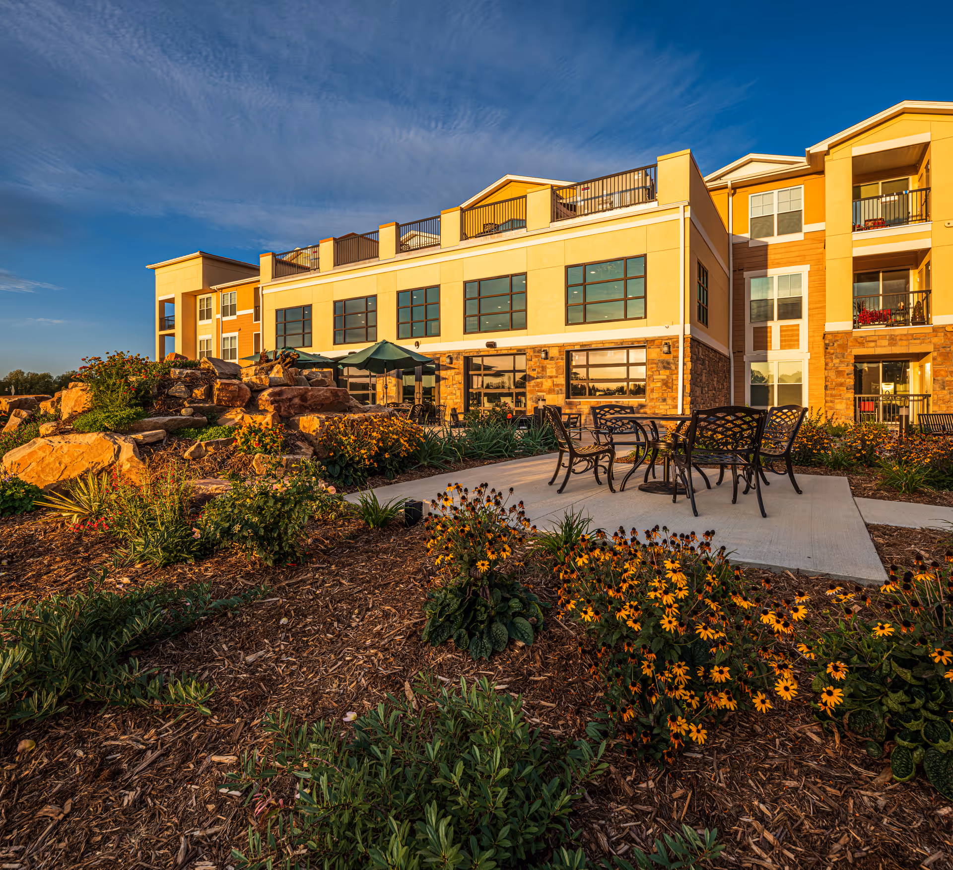 Outdoor patio area at a senior living facility with metal tables and chairs on a concrete slab surrounded by landscaped flower beds and rocks, with a multi-story building in the background under a blue sky.