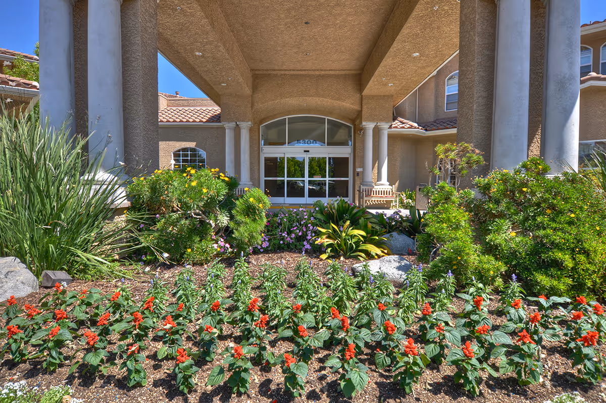 Entrance of a building with large columns and a covered porch, surrounded by well-maintained landscaping including green shrubs and red flowering plants under a clear blue sky.