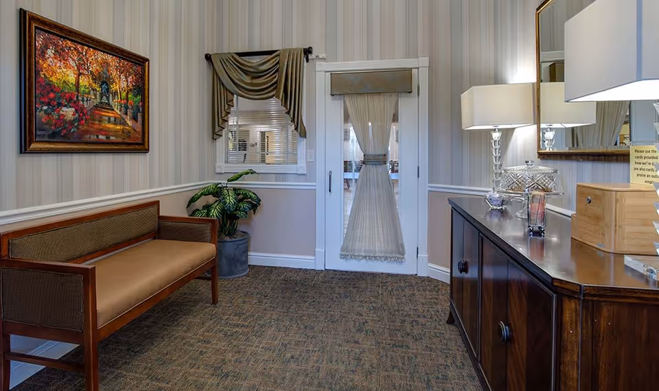 A cozy waiting area with a wooden bench featuring brown cushions, a potted plant in the corner, and a sideboard with a lamp, glass containers, and a wooden box. The walls have striped wallpaper and a framed painting of a colorful garden pathway. A window with draped curtains and a door with sheer curtains are also visible.