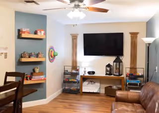 A cozy living room area with a mounted flat-screen TV on the wall, two wooden decorative columns on either side, a wooden console table with decorative lanterns and storage baskets underneath, a brown leather armchair, a ceiling fan with lights, floating shelves with board games and snacks, and a colorful wall decoration shaped like a sombrero.