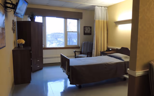 A simple, clean nursing facility bedroom with a single bed covered in a brown blanket, a wooden nightstand with a phone, a striped armchair near a window with a view outside, a wooden cabinet with a small plant and flowers on top, and a wall-mounted TV in the corner.