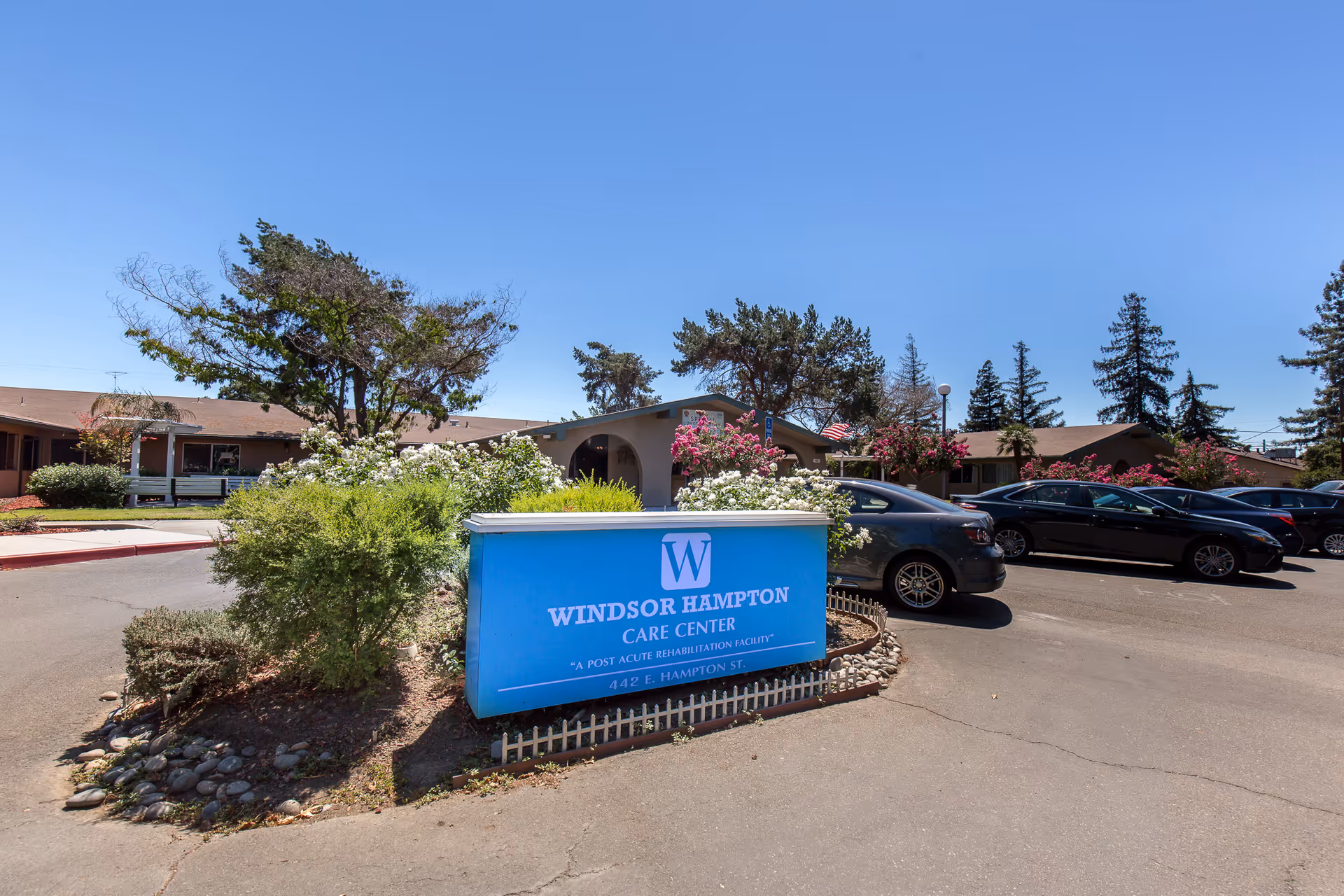 Exterior view of Windsor Hampton Care Center with a blue sign in front displaying the facility name and address. The building is single-story with a parking lot containing several cars. Trees and flowering bushes surround the area under a clear blue sky.