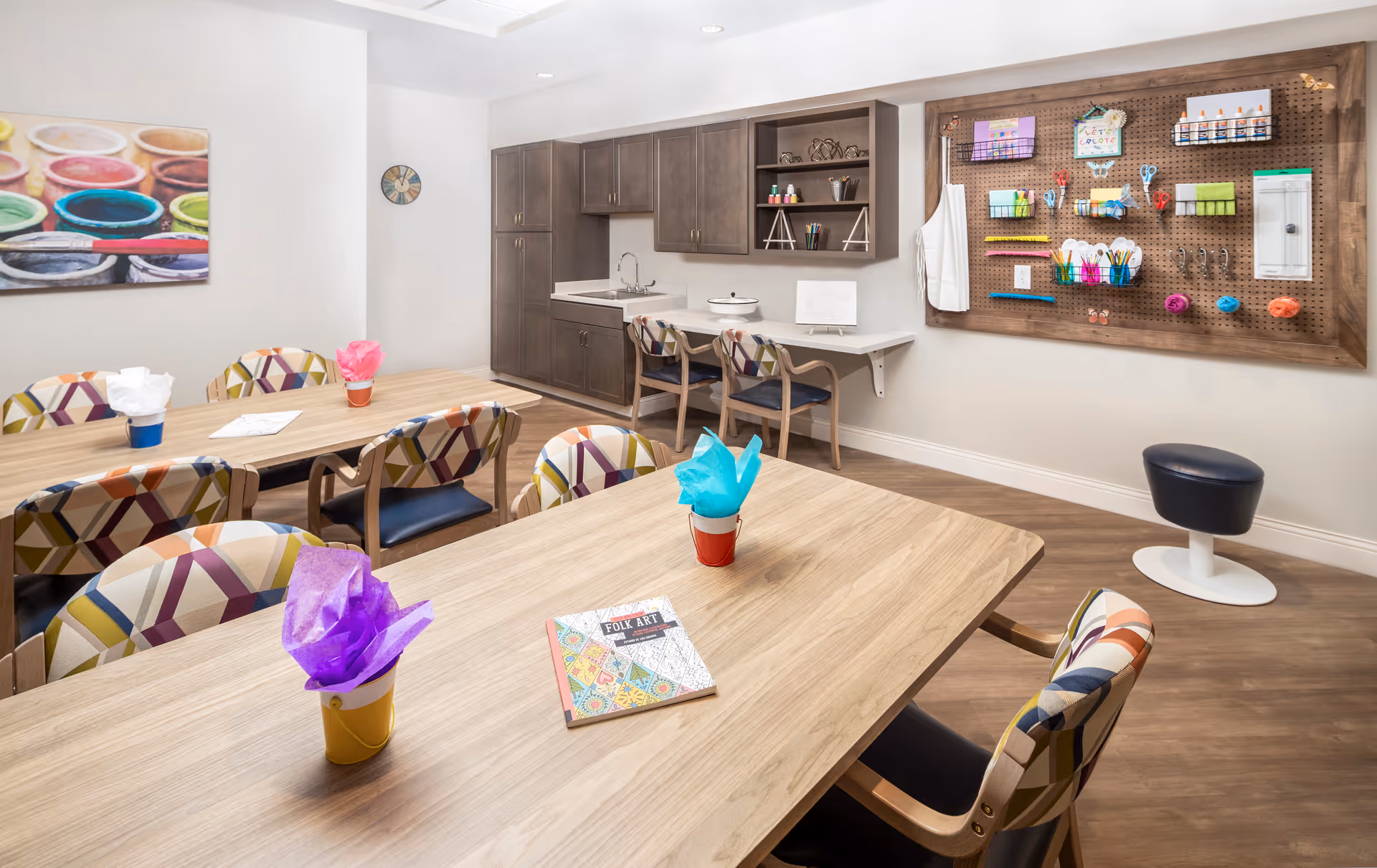 A bright and organized activity room with wooden tables and colorful patterned chairs. On the tables are small buckets with tissue paper and a book titled 'Folk Art'. The back wall features a pegboard with various art supplies such as scissors, tape, and markers. There are cabinets and a small sink area along the back wall, and a colorful painting of paint pots hangs on the left wall.
