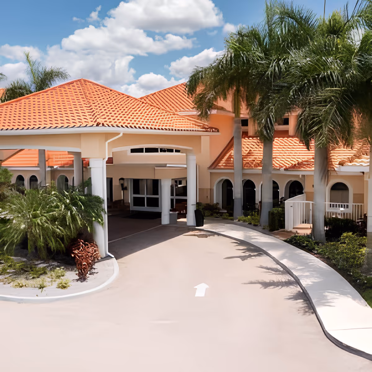 Exterior view of Discovery Village At The Forum showing a building with a red tiled roof, arched windows, palm trees, and a covered driveway entrance under a partly cloudy sky.