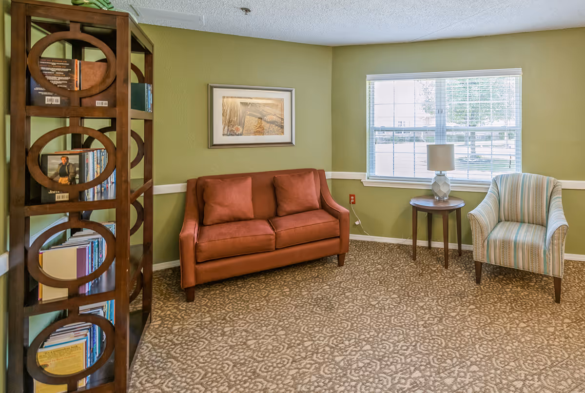 A cozy sitting area with a brown loveseat, a striped armchair, a round wooden side table with a lamp, a bookshelf filled with books, and a window with white blinds letting in natural light. The walls are painted green and the floor is carpeted with a patterned design.
