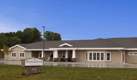 Exterior front view of a single-story assisted living facility building with beige siding, white trim, and a dark roof. There is a covered entrance supported by white columns. A sign in front reads 'Windsor Manor 512'. The surrounding area has green grass and some trees in the background under a clear blue sky.