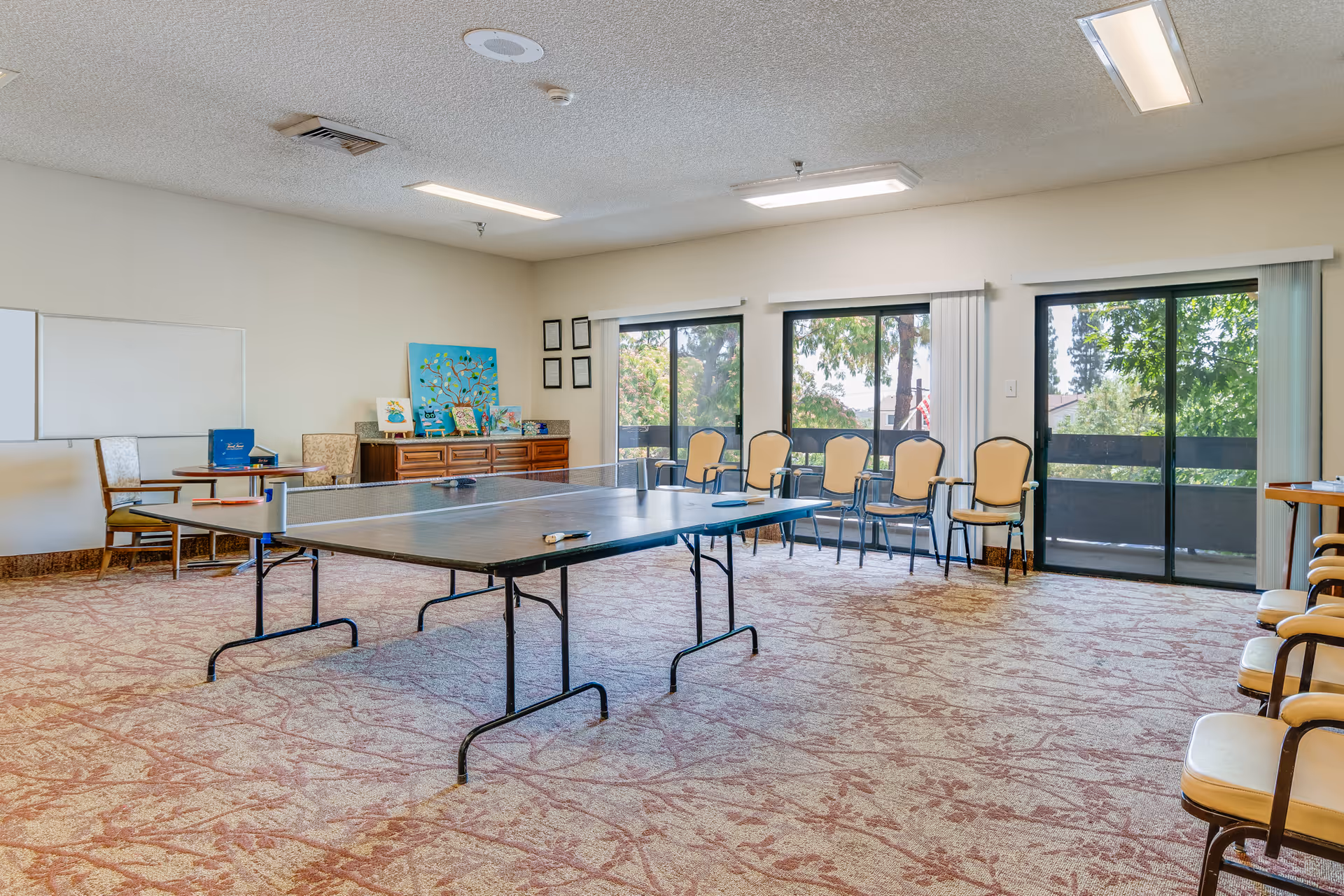 A spacious room with a ping pong table in the center, surrounded by chairs along the walls. The room has large windows and sliding glass doors letting in natural light, with a carpeted floor and ceiling lights. There is a wooden cabinet with decorative items and artwork on the far wall.