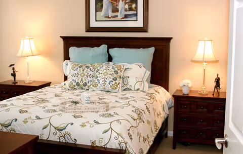A neatly made bed with a floral patterned bedspread and multiple pillows in a bedroom. There are two wooden nightstands on either side of the bed, each with a lamp and decorative items. A framed painting hangs on the wall above the headboard.