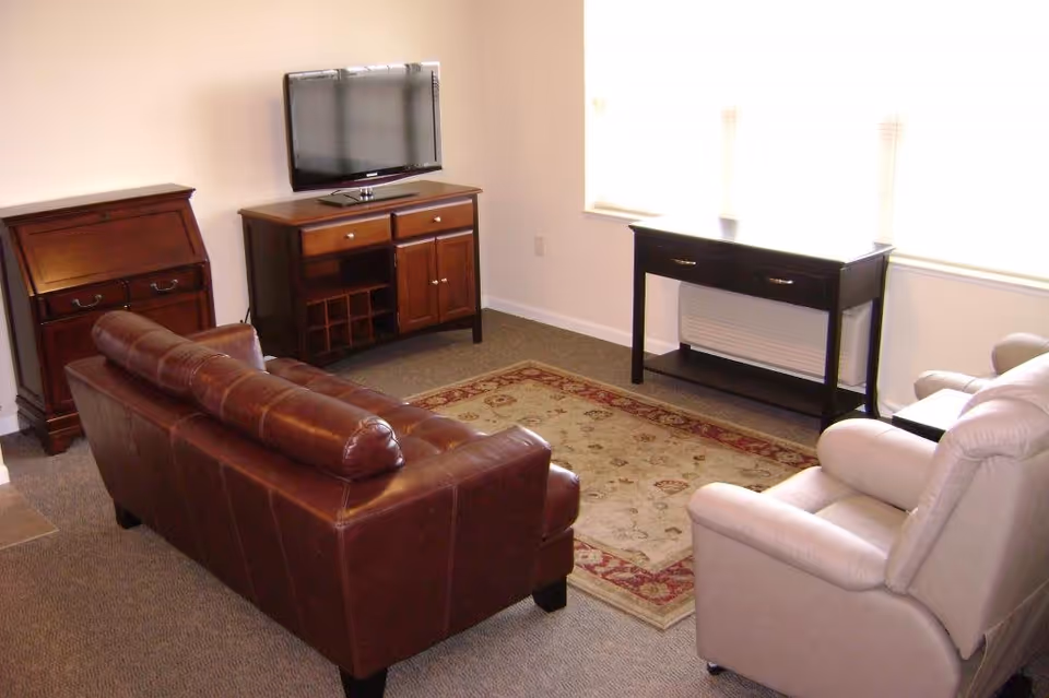 A living room with a brown leather sofa, a beige recliner chair, a wooden TV stand with a flat-screen TV mounted above it, a wooden cabinet, a decorative area rug, and a console table in front of two large windows with blinds.