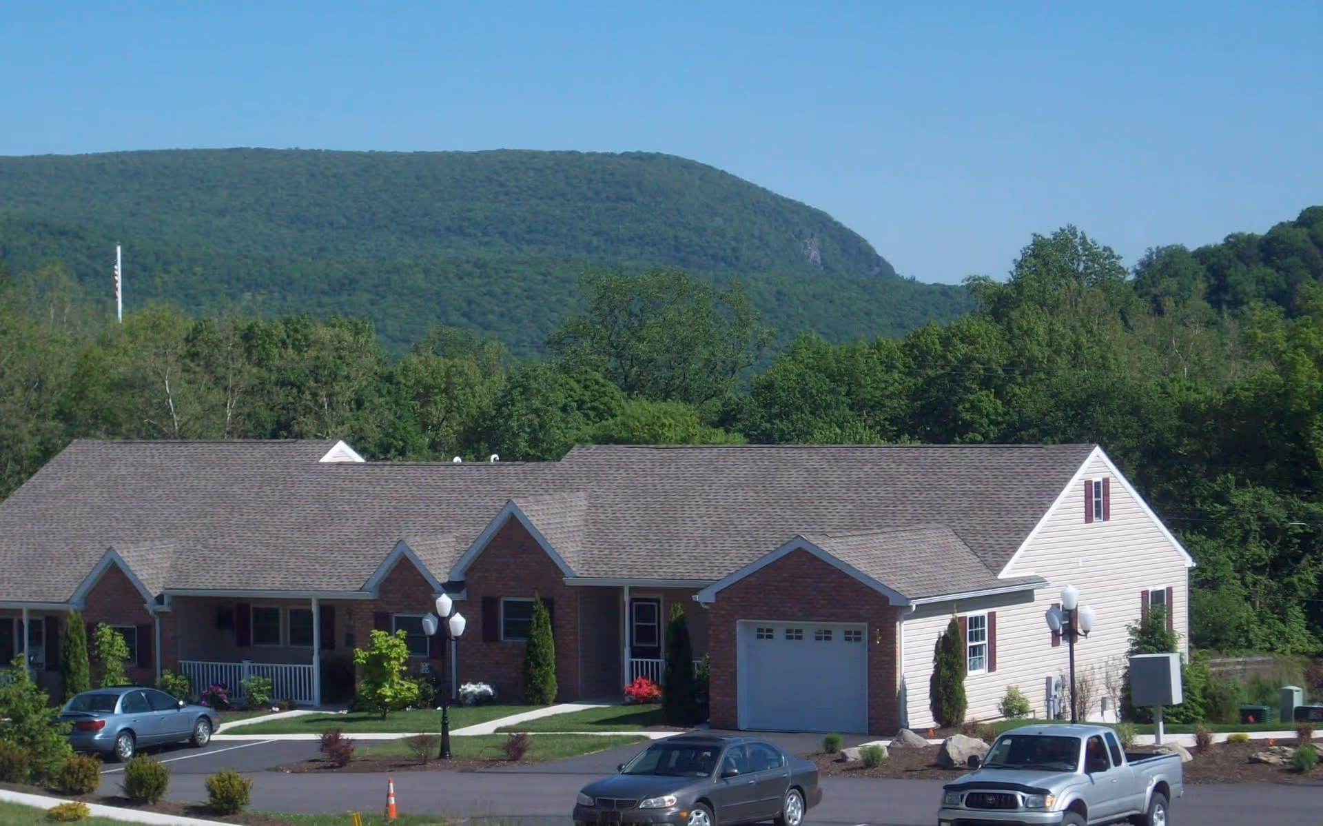 Exterior view of a single-story residential building with brick and siding, surrounded by greenery and trees, with a mountain in the background and several parked cars in the driveway.