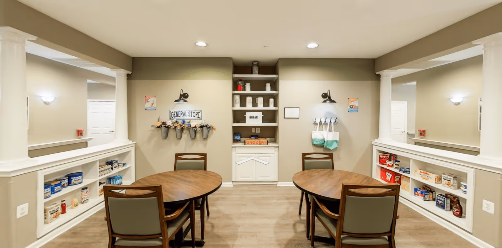 Interior view of a general store area in a senior living facility featuring two round wooden tables with chairs, shelves stocked with grocery items, decorative flower pots on the wall, and a sign that reads 'GENERAL STORE'. The space has beige walls, white trim, and recessed lighting.
