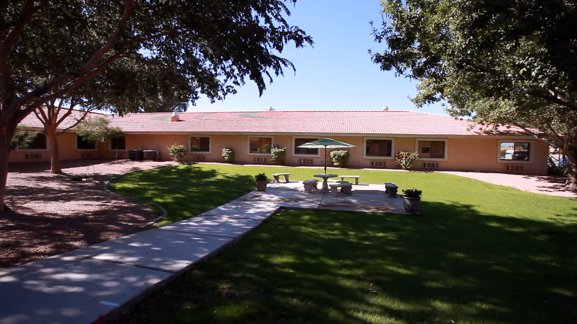 Outdoor courtyard area of Desert Cove Nursing Center with a paved walkway leading to a seating area with a table, benches, and an umbrella. The courtyard is surrounded by a single-story building with windows and shaded by trees.