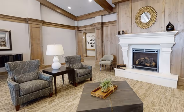 A cozy living room area with two patterned armchairs and a small table with a white lamp between them. There is a modern fireplace with a white mantel and a decorative round mirror above it. A small plant in a wooden tray sits on a dark coffee table in the center. The room features wood paneling on the walls and a beige patterned carpet.