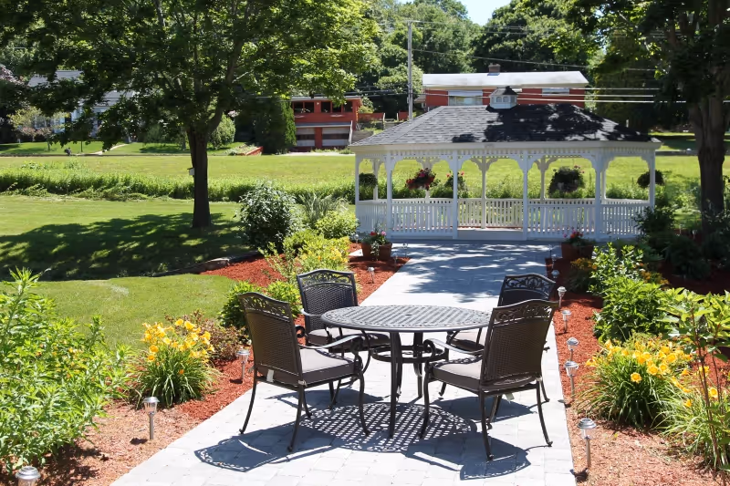 Outdoor garden area with a paved walkway leading to a white gazebo. Four metal chairs with cushions surround a round metal table on the walkway. The garden is landscaped with green bushes, yellow flowers, and mulch beds. Trees and a grassy lawn are visible in the background under a clear sky.