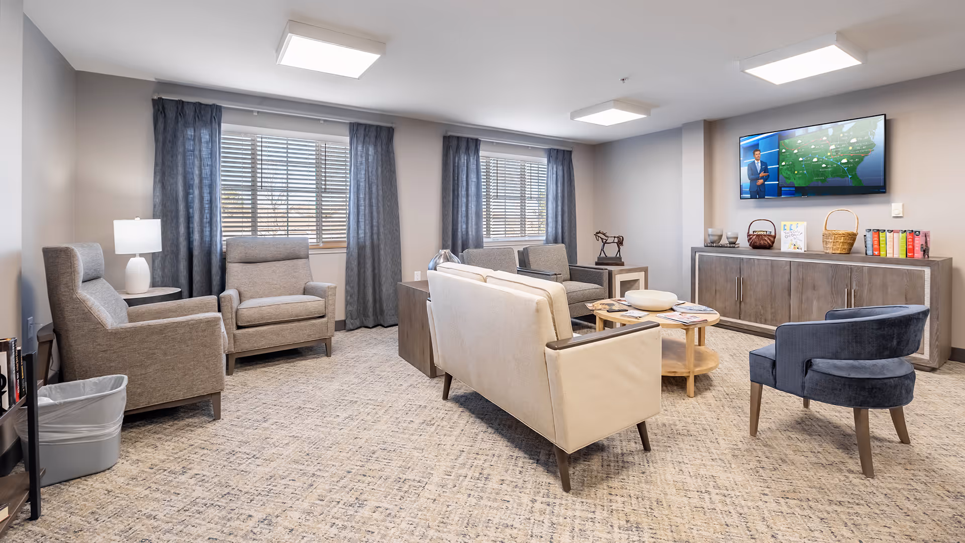 Bright communal lounge with multiple armchairs and sofas arranged around a central coffee table and a wall-mounted TV.
