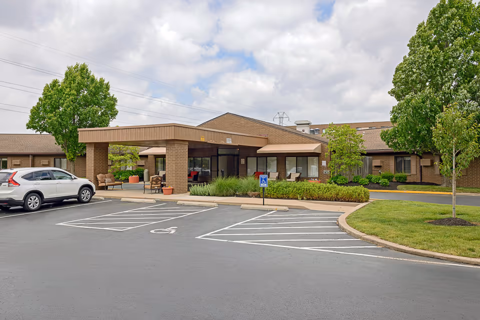 Front entrance of a single-story nursing care center with a covered porte-cochère, outdoor seating, landscaped beds, and adjacent parking with handicap spaces.