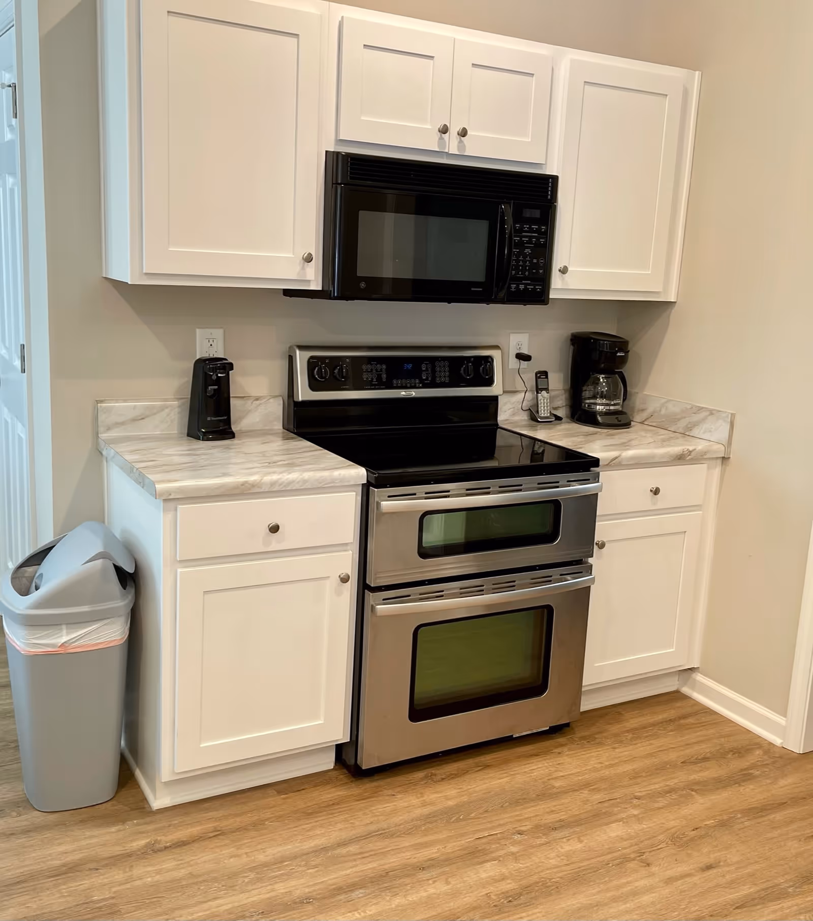 A modern kitchen area with white cabinets, a stainless steel double oven, a black microwave mounted above the oven, a coffee maker, a cordless phone, a black electric can opener, and a gray trash can with a swing lid. The floor is wood, and the countertops have a light marble pattern.