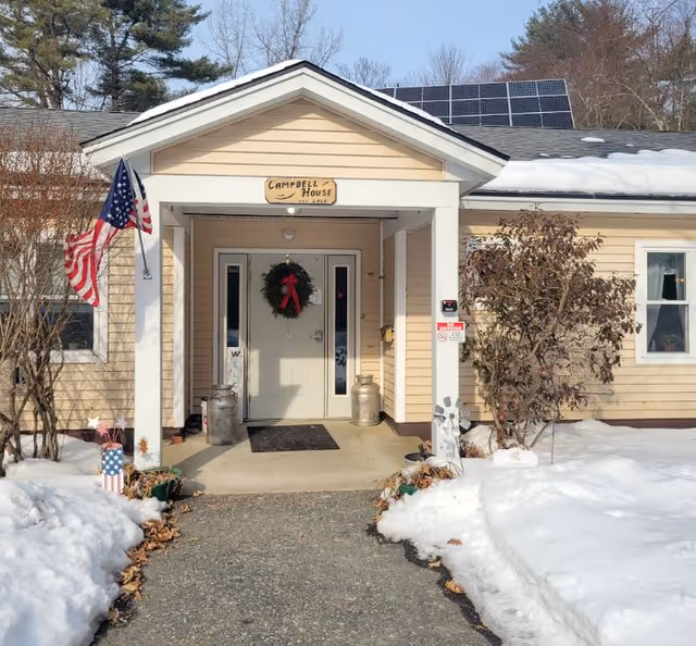 Front entrance of a light-yellow building labeled "Campbell House" with a covered porch, wreath on the door, an American flag, snow along the walkway, and solar panels on the roof.