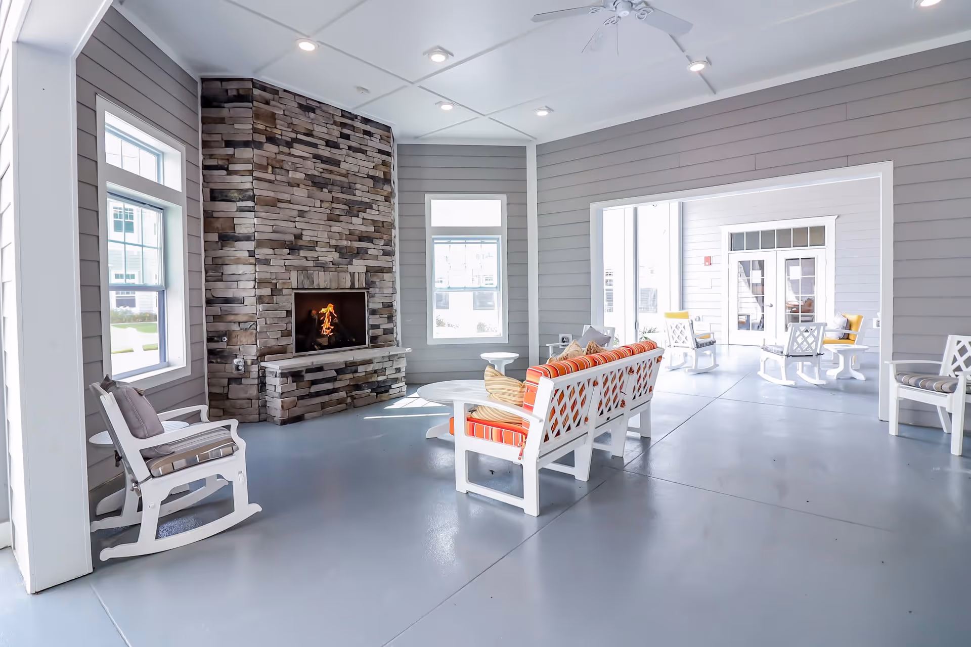 A bright and spacious indoor common area with gray walls and a stone fireplace with a fire burning. The room features white wooden rocking chairs and cushioned benches with orange and beige striped pillows. Large windows allow natural light to fill the space, and there is an open doorway leading to another seating area with similar furniture.