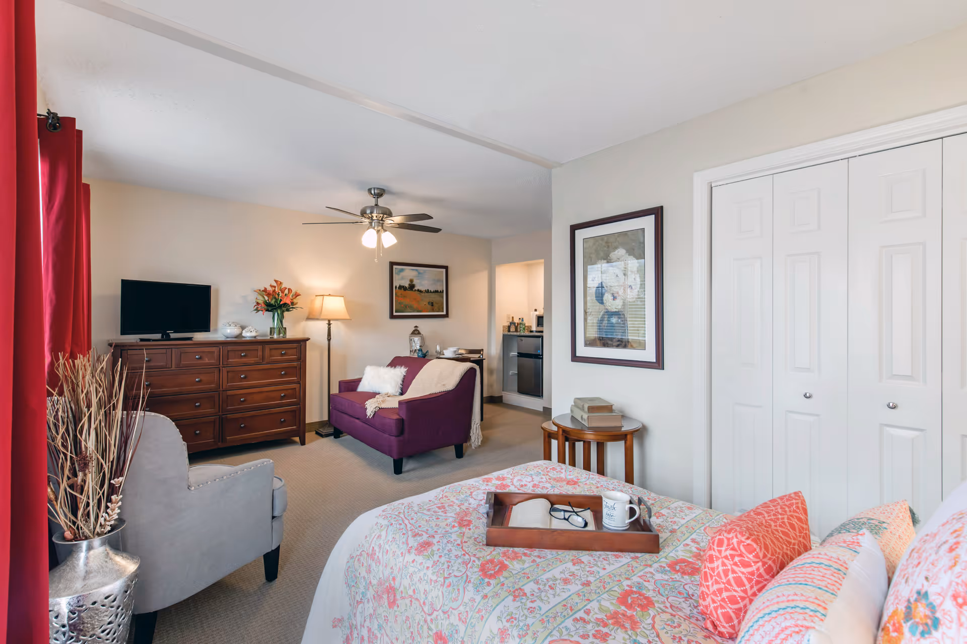 Bright furnished studio-style room with a bed in the foreground, seating area with a purple loveseat and TV on a dresser, ceiling fan, and closet doors.