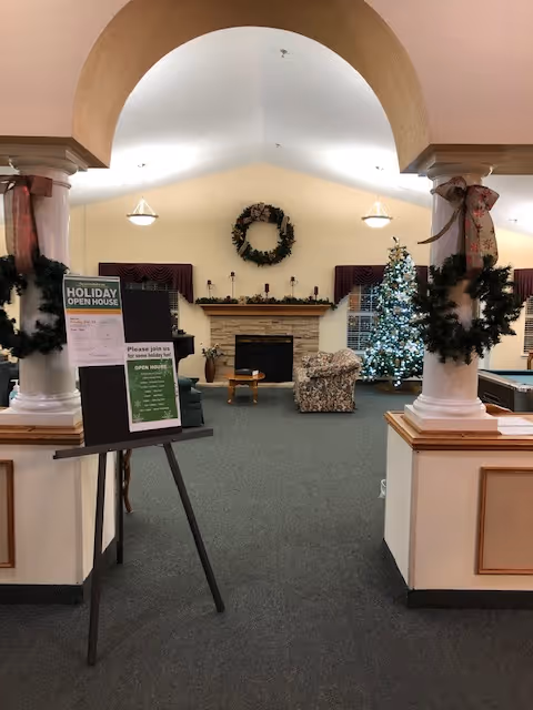 Interior view of a common living area decorated for the holidays with a Christmas tree, wreaths on the walls and columns, a fireplace with a mantel, and a sign on an easel announcing a Holiday Open House event.