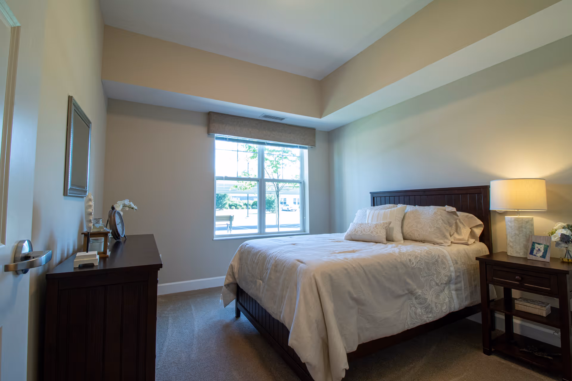A neatly made bed with white and beige bedding in a bedroom with beige walls and carpeted floor. There is a wooden nightstand with a lamp, a framed photo, and a small flower arrangement next to the bed. A wooden dresser with decorative items and a mirror above it is positioned against the opposite wall. A large window with a beige valance allows natural light into the room.