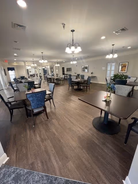 A spacious dining room with multiple dark wood tables and cushioned chairs arranged neatly. The room has wood flooring, light-colored walls, and several ceiling light fixtures. There are framed pictures on the walls and some plants placed around the room, creating a welcoming atmosphere.