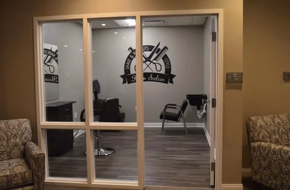 Interior view of a beauty salon in an assisted living facility, showing two salon chairs, a hair washing station, and a wall decal with scissors and combs that reads 'Shave & Haircuts Style Hair Salon'. The room is visible through a large glass window and door, with two patterned armchairs outside the salon.