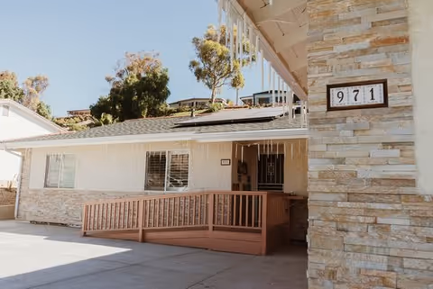 Exterior view of a single-story building with a stone facade and a wooden ramp leading to the entrance. The building number 971 is displayed on the wall. There are icicle-style decorations hanging from the roof edge and trees in the background under a clear sky.
