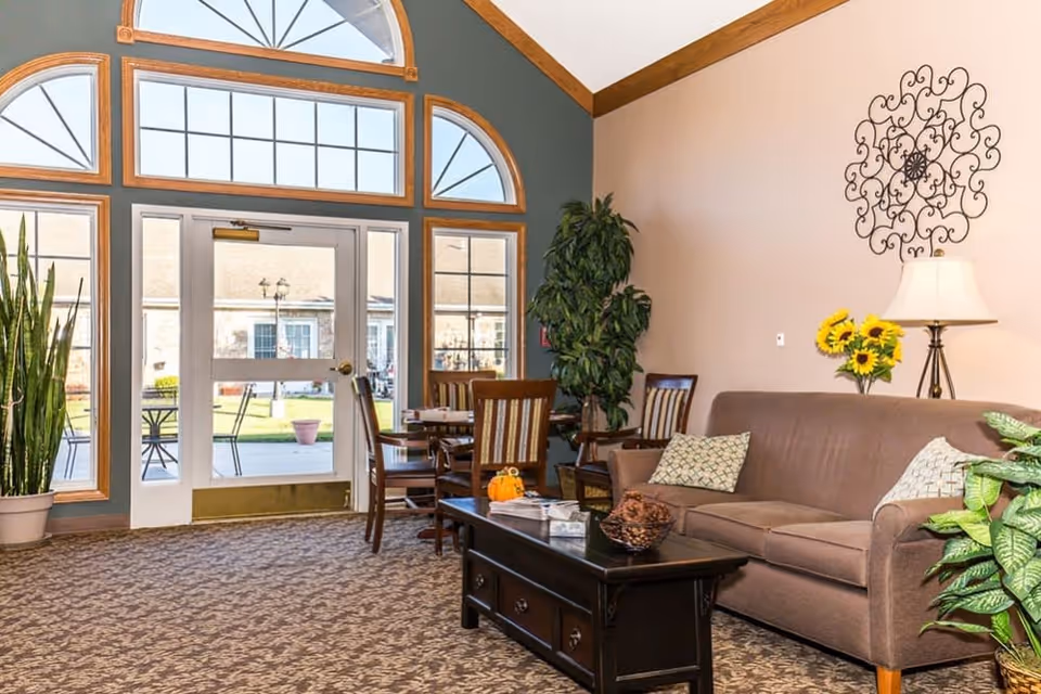 A cozy living room area with a brown sofa adorned with patterned pillows, a dark wooden coffee table with decorative items, and a side table with a lamp and a vase of sunflowers. There are several green plants around the room, and a large window and glass door letting in natural light, showing an outdoor patio with tables and chairs.