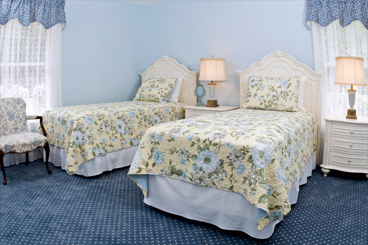 Two twin beds with floral quilts in a light-blue bedroom with matching nightstands, lamps, and a patterned chair by lace-curtained windows.