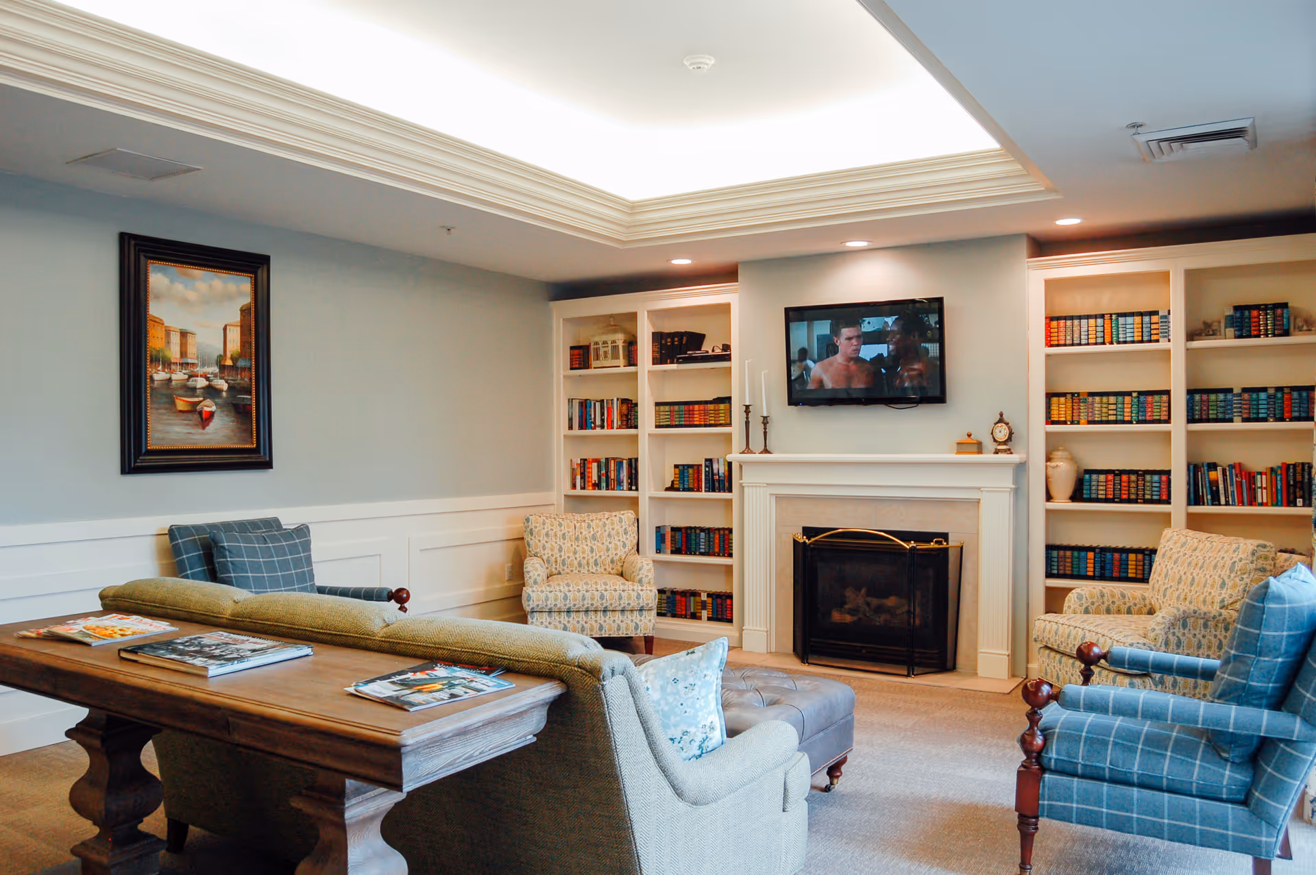 A cozy living room with a fireplace, built-in bookshelves filled with books, a wall-mounted TV showing a scene from a movie, and comfortable seating including armchairs and a sofa. A wooden table behind the sofa holds magazines. The room has soft lighting and a framed painting on the wall.
