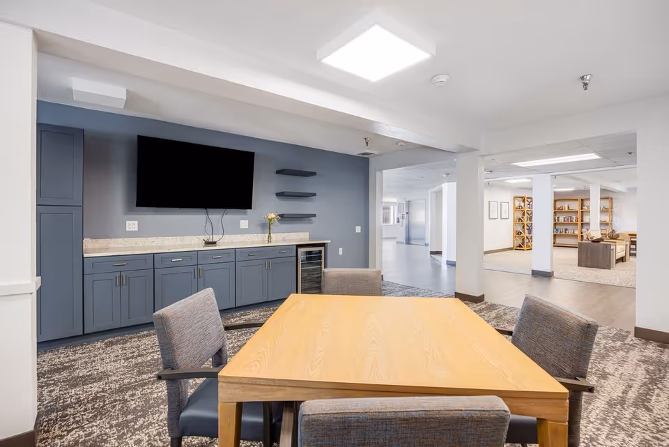 A modern interior room with a wooden table surrounded by four gray upholstered chairs. Against the wall is a blue cabinet with a granite countertop, a mounted flat-screen TV, and three floating shelves. The room opens into a hallway and a lounge area with bookshelves and seating.