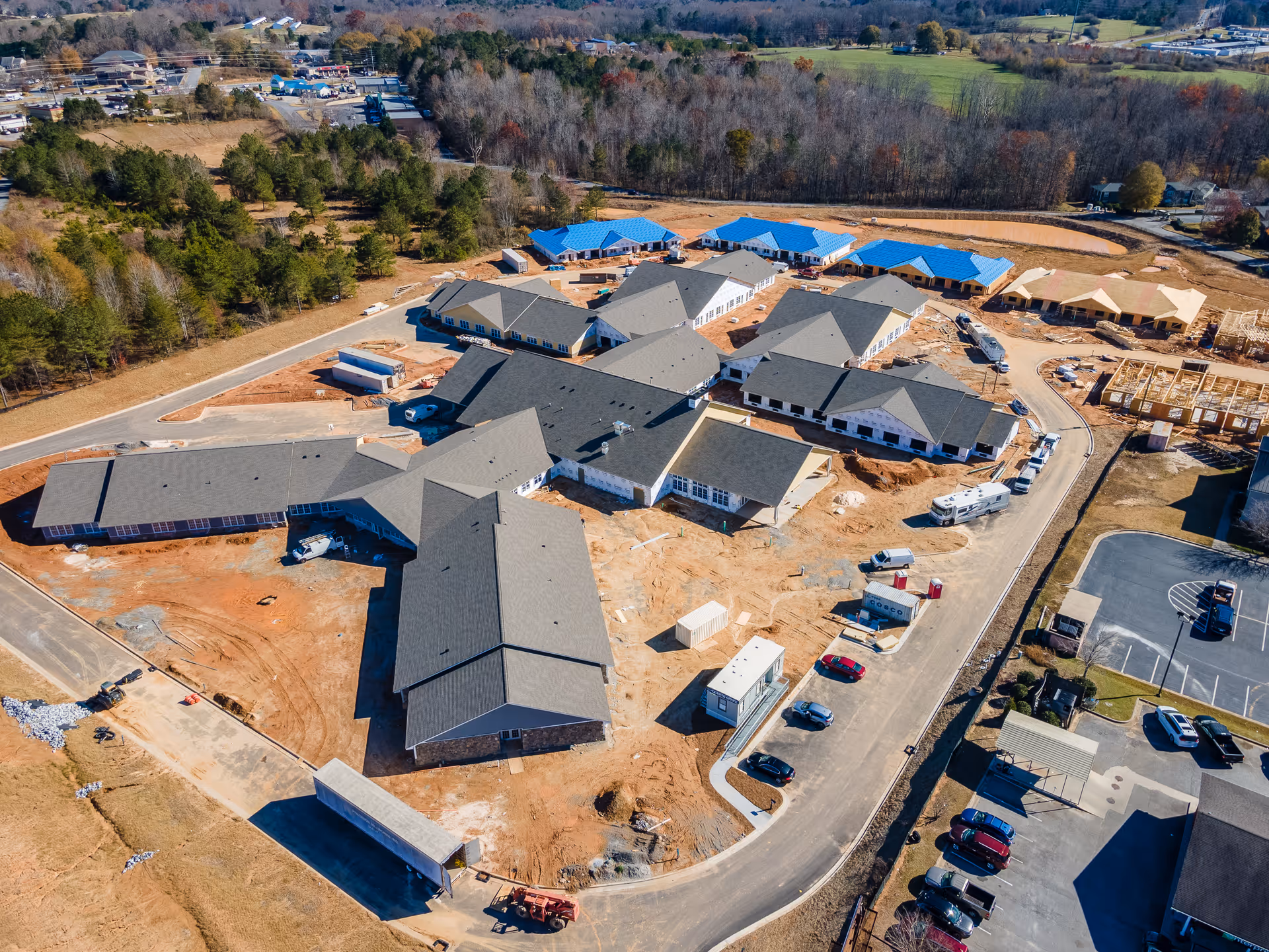 Aerial view of Manor Lake Assisted Living, Memory Care, and Independent Living facility under construction in Dawsonville, showing multiple buildings with gray and blue roofs, construction equipment, vehicles, and surrounding wooded area.