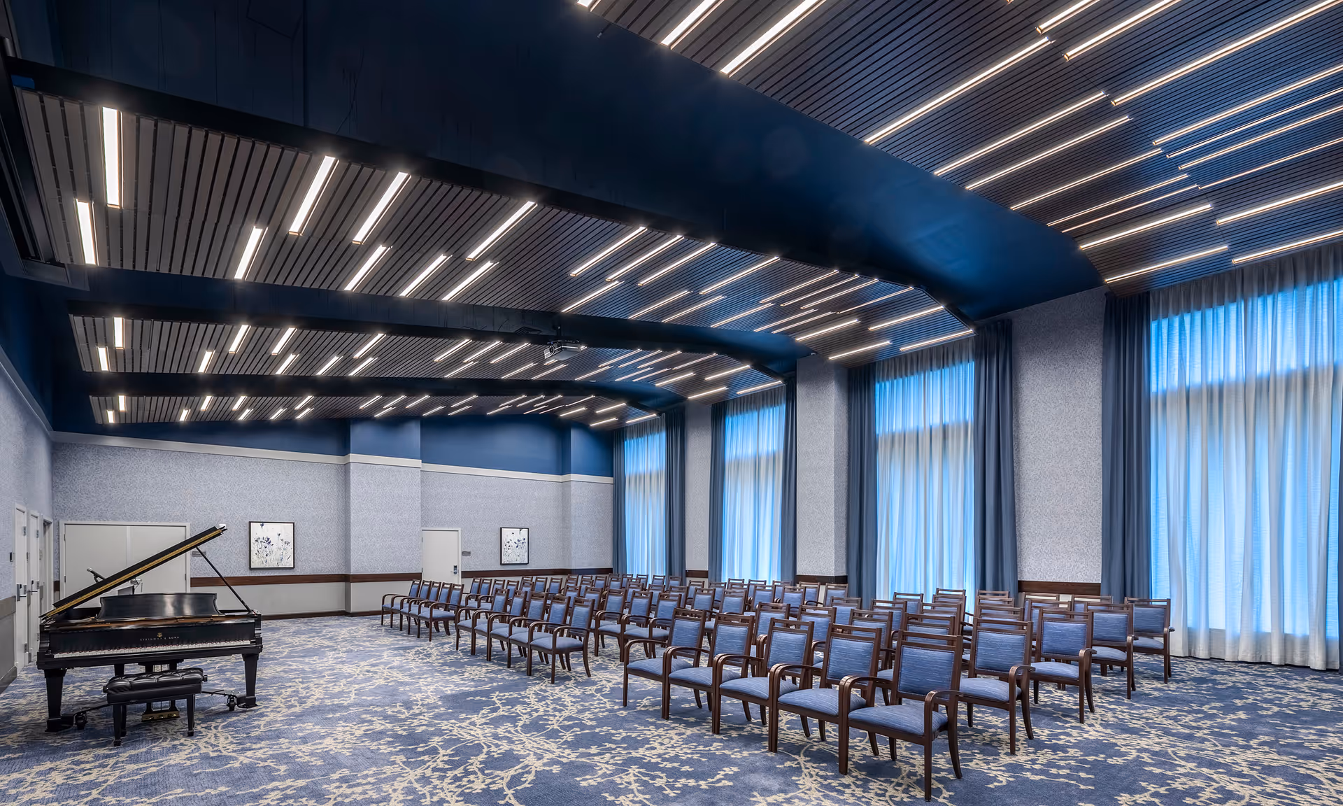 Spacious event room with rows of chairs, a grand piano at the front, blue carpeting and tall curtained windows under a modern lit ceiling.