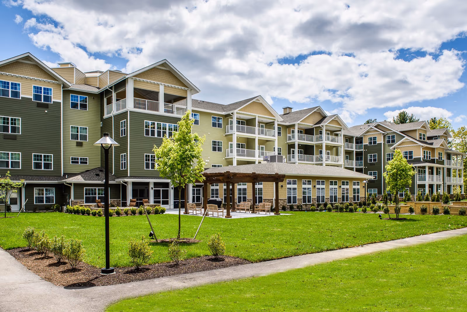 Exterior view of a multi-story senior living facility with green and beige siding, multiple balconies, a well-maintained lawn, small trees, shrubs, a paved walkway, and a wooden pergola with outdoor seating under a partly cloudy sky.