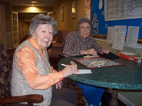 Two elderly women sitting at a round table playing a board game in a common area of a senior living facility. One woman is wearing an orange shirt and a beige vest, while the other is wearing a striped shirt. Behind them is a bulletin board with various notices and a hallway leading to other rooms.