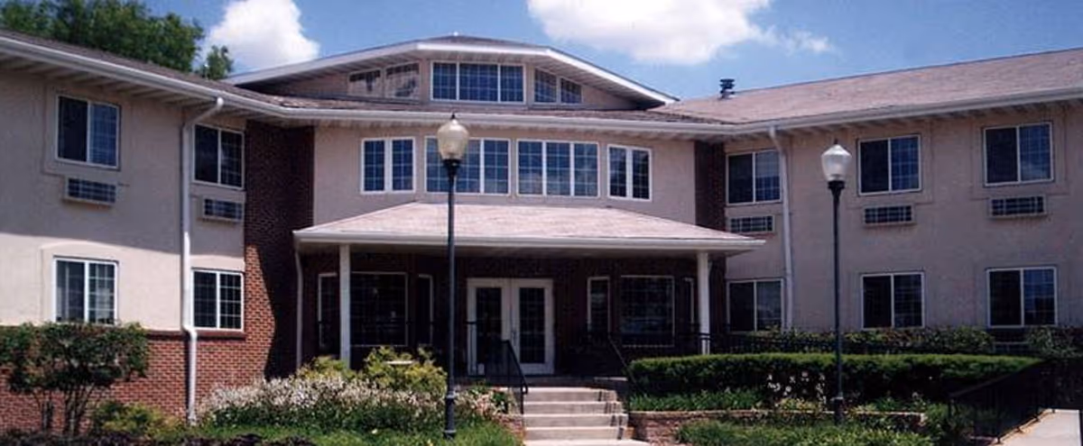Front exterior view of a two-story assisted living facility building with multiple windows, a covered entrance with steps leading up, two lamp posts, and landscaped bushes and flowers in front.